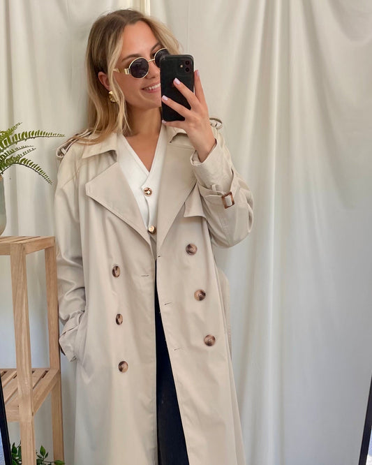 Trench beige coupe longue avec épaules structurées et une ceinture à nouer. Doublé intégralement. 