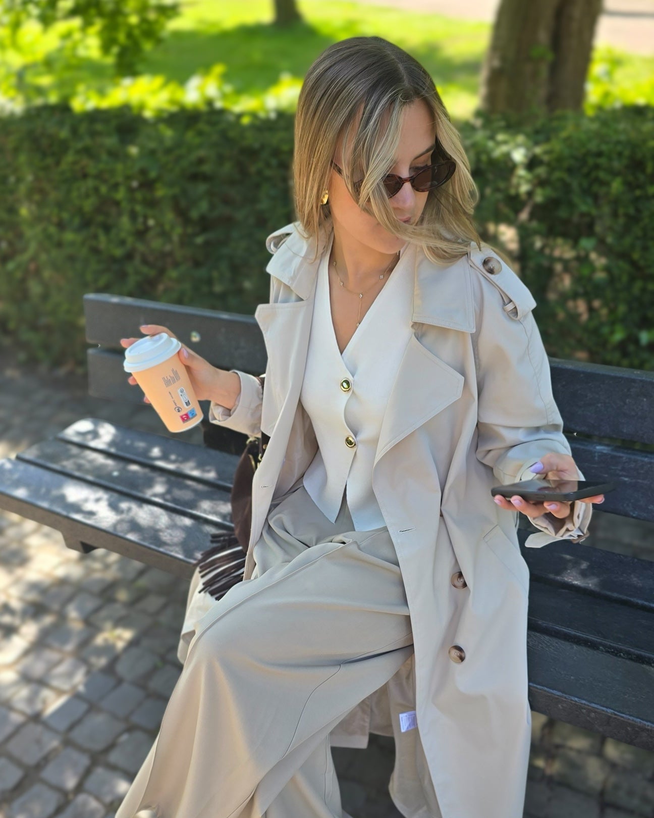 Top sans manches en maille douce en blanc. Encolure en V croisée avec deux boutons dorés sur le devant. Porté avec un trench beige et un pantalon tailleur beige. 