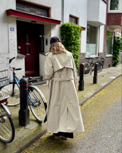 Trench beige coupe longue avec épaules structurées et une ceinture à nouer. Doublé intégralement. 