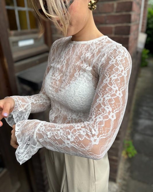 Blouse blanche en dentelles légère et transparente. Coupe moulante avec des longues manches évasées et une dentelle florale. 