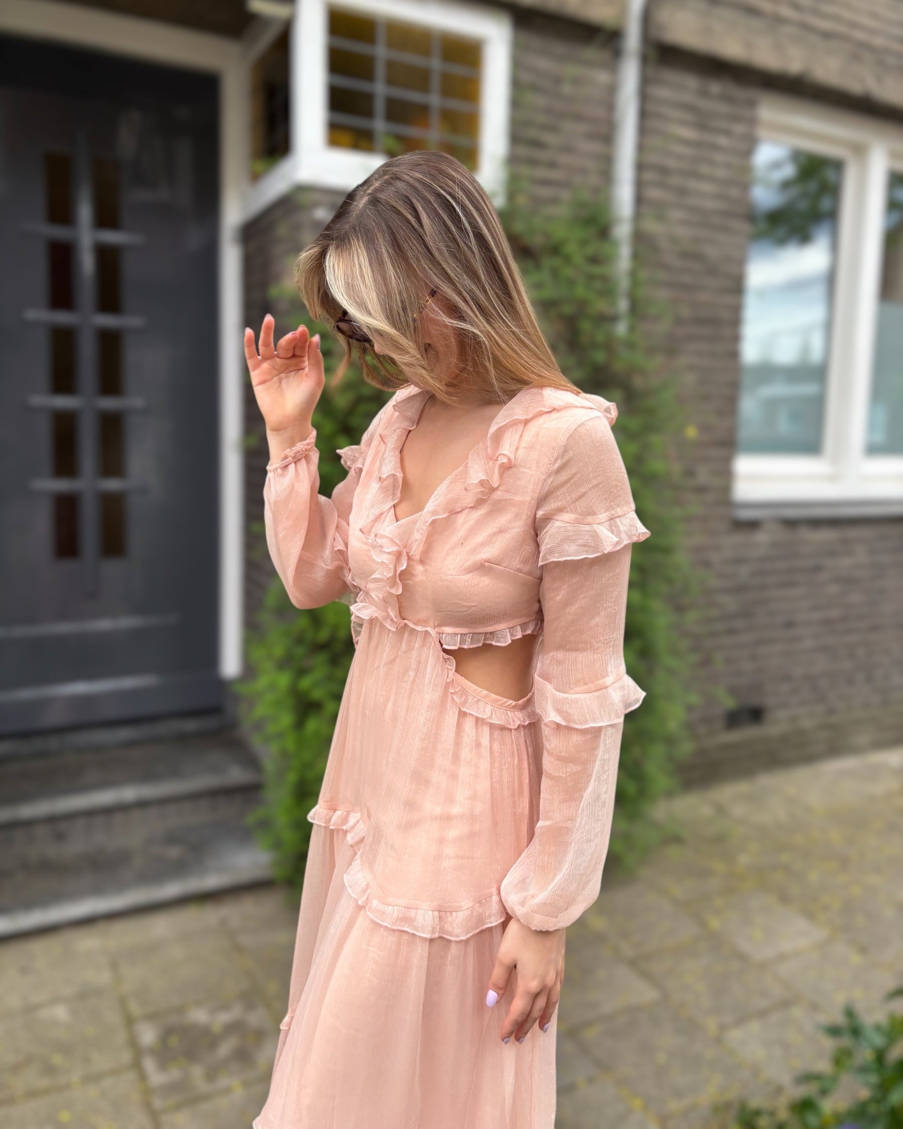 Robe rose longue doublée jusqu’aux cuisses. Décolleté en V avec petits volants. Détails ajourés sur les côtés. 
