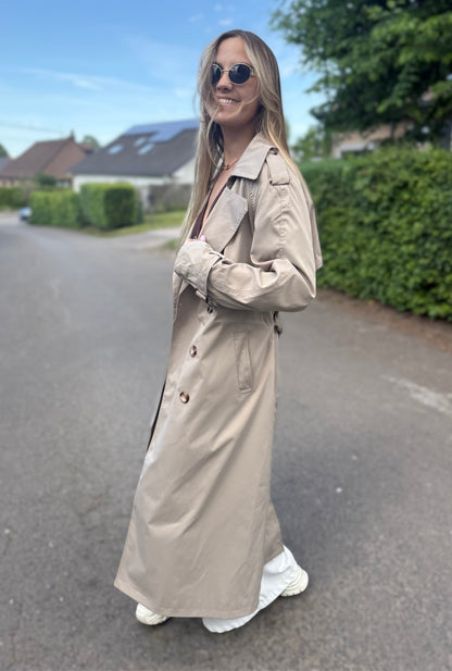 Trench taupe coupe longue avec épaules structurées et une ceinture à nouer. Doublé intégralement. 
